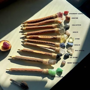 May include: A collection of handcrafted pencils with natural wood shafts, each adorned with a different gemstone. Gemstones include Strawberry Quartz, Sunstone, Chalcopyrite, Howlite, Citrine, Sodalite, Rock Crystal, Jet Stone, Smoky Quartz, and Green Fluorite. The word "LUCKY" is carved on one pencil.