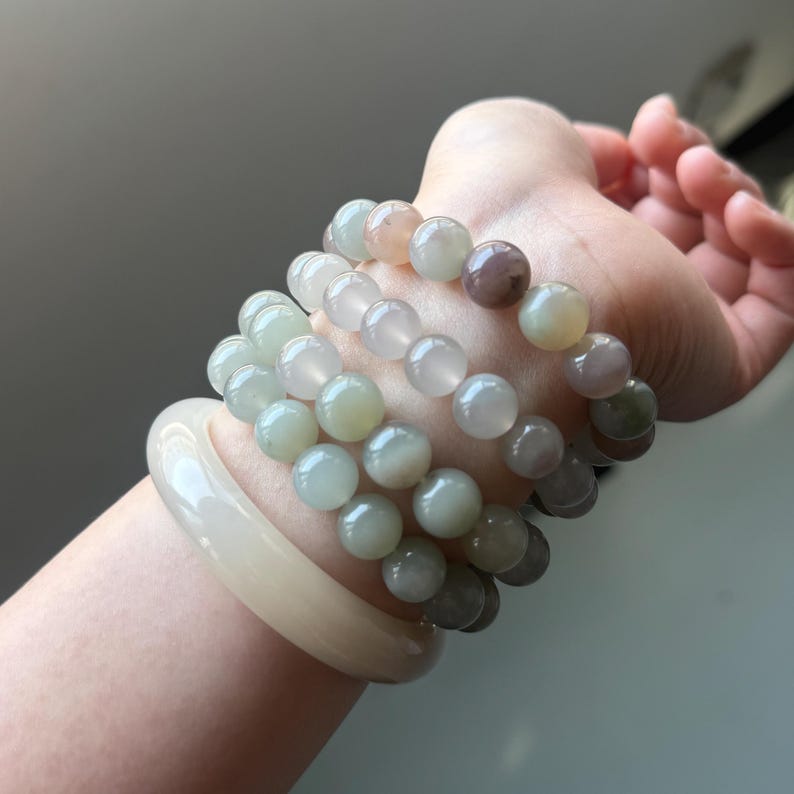 May include: A hand displays a collection of jade bracelets and beaded bracelets. The bracelets feature a variety of colours, including pale green, white, and purple. The jade bracelet is a solid, light cream colour.