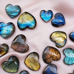 May include: A collection of heart-shaped gemstones in various colors, including blue, gold, and brown. The polished stones have a glossy finish and are arranged on a pink fabric background. These decorative stones are ideal for jewelry making or collecting.