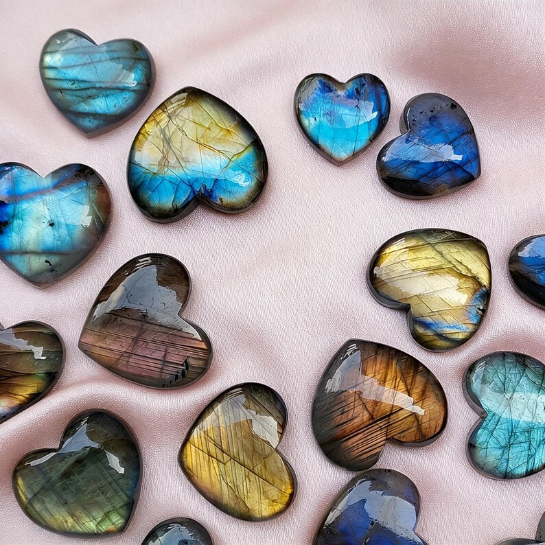 May include: A collection of heart-shaped gemstones in various colors, including blue, gold, and brown. The polished stones have a glossy finish and are arranged on a pink fabric background. These decorative stones are ideal for jewelry making or collecting.
