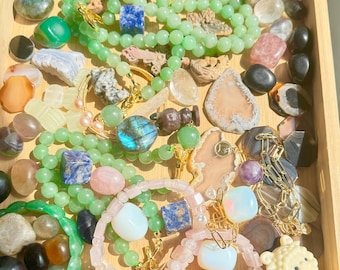 May include: A wooden tray filled with a variety of gemstones and beaded bracelets. The bracelets are in shades of green and pink, with gold accents. The gemstones are in various colors and shapes, including blue, pink, and orange.