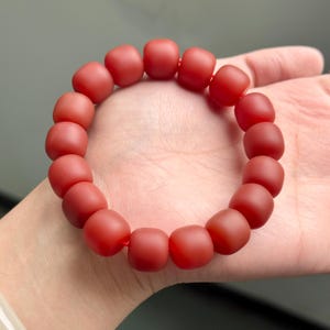Puede incluir: Una pulsera de cuentas rojas con cuentas cuadradas redondeadas. Las cuentas están ensartadas en una cuerda roja, formando una forma circular. La pulsera se muestra en la mano de una persona.