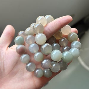May include: A bracelet made of smooth, spherical gemstone beads in shades of pale green, grey, and cream. The beads are strung together. The bracelet is held in a hand.