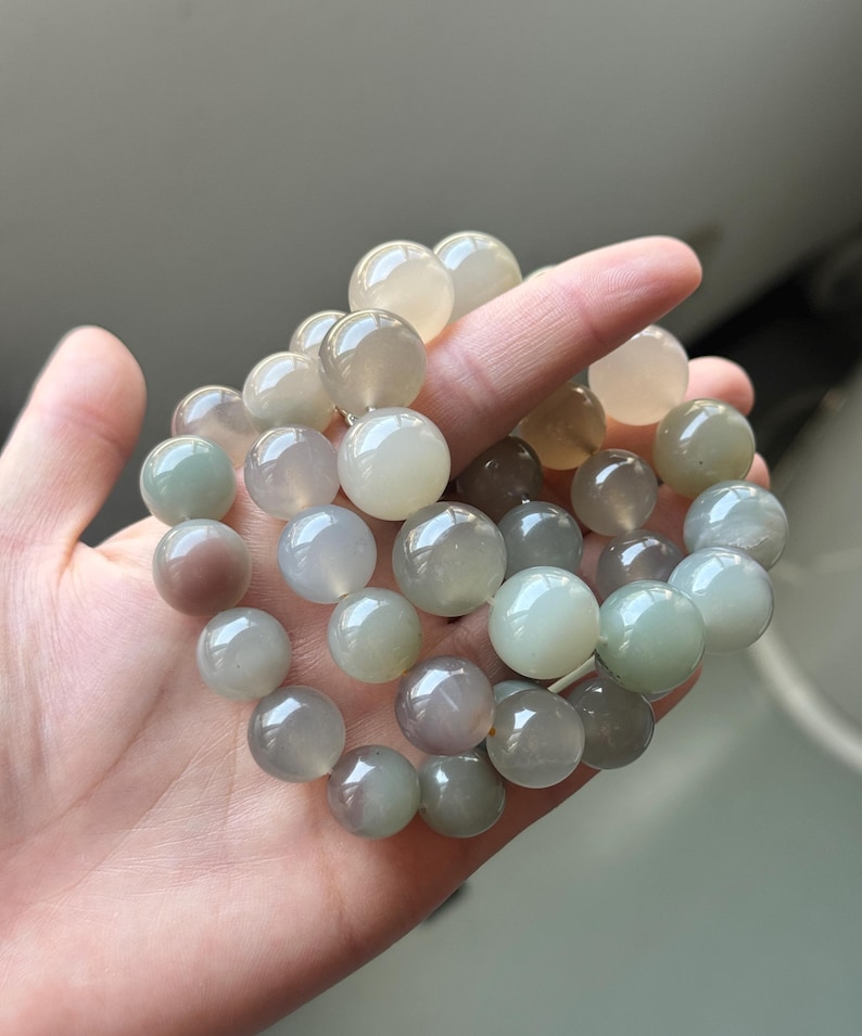May include: A bracelet made of smooth, spherical gemstone beads in shades of pale green, grey, and cream. The beads are strung together. The bracelet is held in a hand.