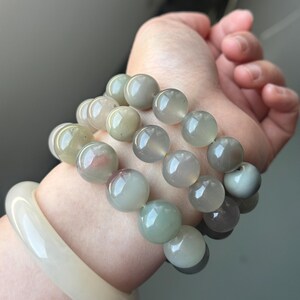 May include: Three beaded bracelets with large, round, translucent beads in shades of green, grey, and pink. A cream-coloured bangle is also visible. The bracelets are worn together, showcasing the varying colours and textures of the beads.