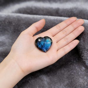 May include: A heart-shaped labradorite gemstone with iridescent blue and teal flashes. The polished stone is held in an open hand, showcasing its natural beauty. The gemstone is approximately 2 inches wide.