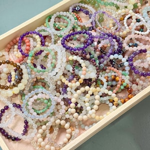 May include: A wooden tray filled with a variety of beaded bracelets. The bracelets are made of different colored stones, including purple, green, pink, and clear beads. The beads are strung together to form a circular shape.