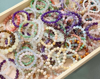 May include: A wooden tray filled with a variety of beaded bracelets. The bracelets are made of different colored stones, including purple, green, pink, and clear beads. The beads are strung together to form a circular shape.