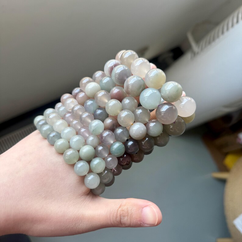 May include: Several beaded bracelets in shades of green, purple, and cream. The bracelets are made of round beads and are stacked together. The beads have a glossy finish and are strung on an elastic cord.