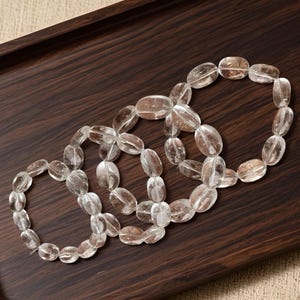 May include: Three clear quartz crystal bracelets with oval-shaped beads are arranged on a dark brown wooden tray. The bracelets are linked together, showcasing the natural clarity and texture of the stones.