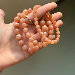 May include: Three strands of peach-colored gemstone bead bracelets. The beads are round and vary in size. A silver ring is visible on a finger. The bracelets are held in a hand against a blurred background.