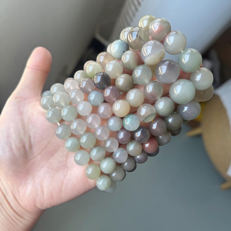 May include: A collection of beaded bracelets featuring a range of pastel-coloured beads, including green, pink, and beige. The round beads are made of a smooth, polished material. The bracelets are held in a hand.
