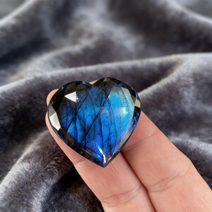 May include: A faceted, heart-shaped labradorite gemstone. The stone displays a vibrant blue and iridescent flash, with dark inclusions. The gemstone is held in a hand against a soft, gray background. The heart shape is clearly defined with multiple facets.