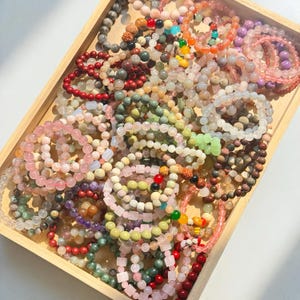 Puede incluir: Una bandeja de madera llena de pulseras de cuentas de varios colores. Las pulseras presentan una gama de tonos, incluyendo rosa, verde, rojo y marrón. Las cuentas están hechas de diferentes materiales y tamaños, creando una diversa colección de joyas.