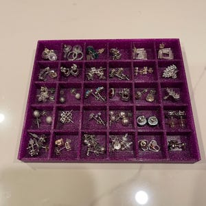 May include: A purple, divided tray filled with various pairs of earrings. The earrings are silver-toned and feature diverse designs, including studs, hoops, and dangling styles. The tray is on a white surface.
