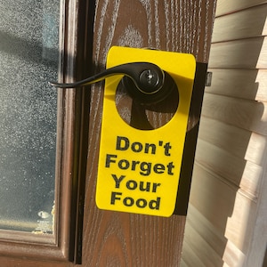 May include: A yellow door hanger with the text "Don't Forget Your Food" in black letters. The door hanger is hanging on a brown door with a black handle.