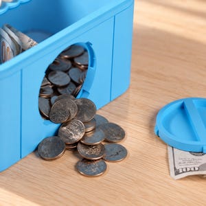 May include: A bright blue plastic piggy bank shaped like a building block with a circular opening on the side. Silver coins are spilling out of the opening onto the surface. A matching blue circular lid sits nearby next to a folded hundred dollar bill.