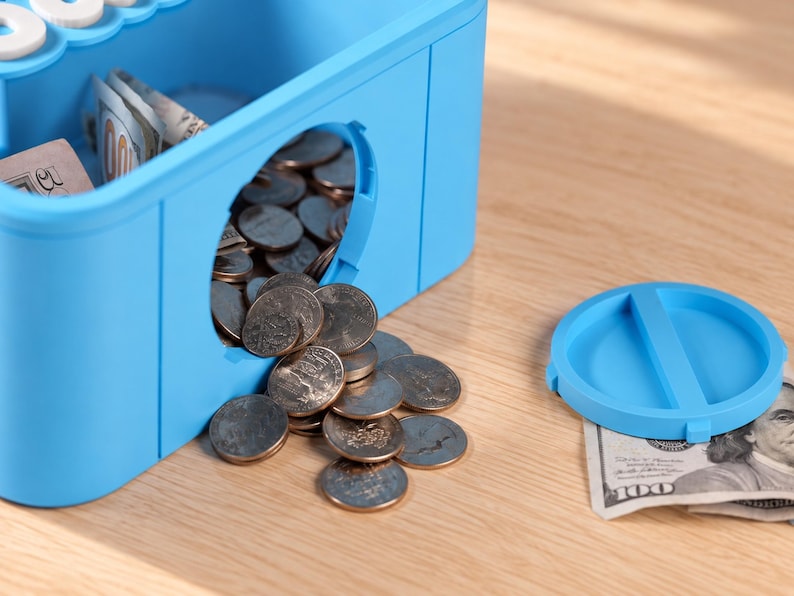 May include: A bright blue plastic piggy bank shaped like a building block with a circular opening on the side. Silver coins are spilling out of the opening onto the surface. A matching blue circular lid sits nearby next to a folded hundred dollar bill.