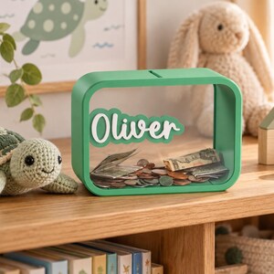 May include: A rectangular green piggy bank with a clear front panel featuring the name Oliver in white script lettering. The bank contains a collection of assorted metal coins and folded paper currency visible through the transparent front surface.
