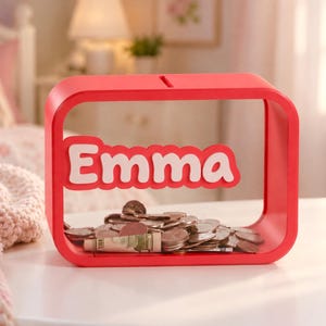 May include: A rectangular red piggy bank with rounded corners and a clear front panel. The name Emma is written in white, raised letters across the center. The interior contains a collection of assorted metal coins and a rolled-up paper bill.