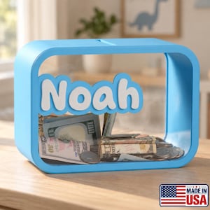 May include: A light blue rectangular piggy bank with rounded corners and a clear front panel. The name Noah is displayed in white, raised lettering across the center. The interior contains various United States paper currency and metal coins.