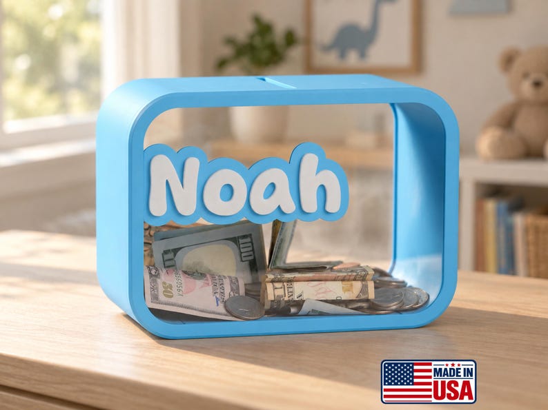 May include: A light blue rectangular piggy bank with rounded corners and a clear front panel. The name Noah is displayed in white, raised lettering across the center. The interior contains various United States paper currency and metal coins.