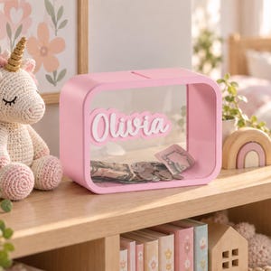 May include: A rectangular pink piggy bank with a clear front panel featuring the name Olivia in white script lettering. The bank has a slot on the top for inserting coins and paper currency, which are visible inside the transparent container.