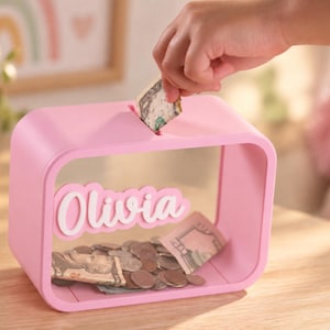 May include: A rectangular pink piggy bank with a clear front panel and a slot on top for inserting money. The name Olivia is written in white script lettering on the front. The interior contains various metal coins and paper currency.