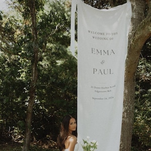 Op de afbeelding: Een bruid in een witte strapless trouwjurk houdt een boeket witte bloemen vast. Een spandoek op een boom leest "WELCOME TO THE WEDDING OF EMMA & PAUL" met de datum 14 september 2024.
