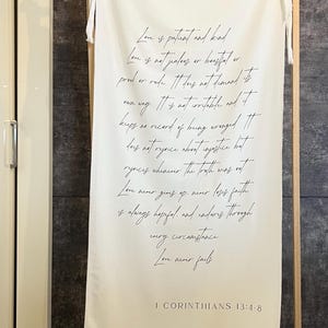 Puede incluir: Una pancarta de tela blanca con escritura negra, exhibida en un marco de madera. El texto incluye un pasaje de 1 Corintios 13:4-8 sobre el amor. La pancarta está colgada en interiores.