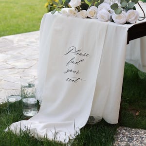 Peut inclure: Une table décorée pour un événement, avec une nappe blanche portant l'inscription "Please find your seat". La table est ornée de roses blanches, de verdure et d'un chandelier doré. Deux vases en verre transparent avec de l'eau et des bougies sont au premier plan.
