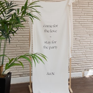 Könnte beinhalten: Ein weißes Stoffbanner mit dem Text "come for the love - stay for the party" in schwarzer Schrift. Das Banner wird auf einem Holzrahmen angezeigt, mit den Initialen "A&N" unten. Eine grüne Pflanze befindet sich im Vordergrund.