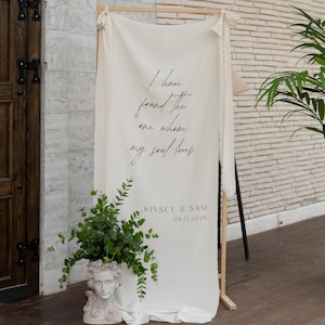 Può includere: Un fondale in tessuto color crema con la scritta "I have found the one whom my soul loves" in elegante corsivo. Sotto, si legge "KINSEY & SAM 09.13.2025". Un vaso decorativo con verde alla base.