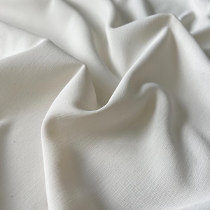 May include: Close-up of white fabric with visible texture and folds. The fabric appears to be a solid, off-white color, with subtle variations in tone due to the light and shadows. The material has a slight sheen.