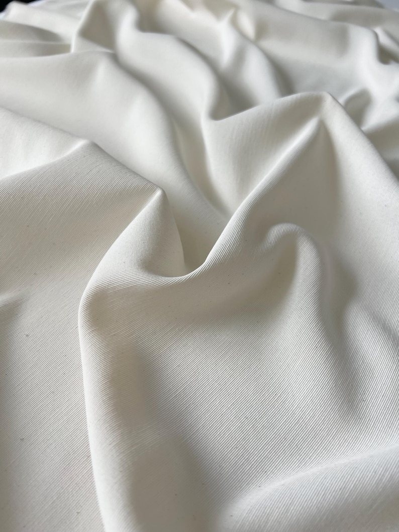 May include: Close-up of white fabric with visible texture and folds. The fabric appears to be a solid, off-white color, with subtle variations in tone due to the light and shadows. The material has a slight sheen.