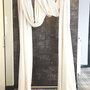 May include: A wooden frame draped with flowing, off-white fabric, creating an archway. The backdrop is a textured, dark gray wall, with a light fixture above. The floor has a plaid pattern.