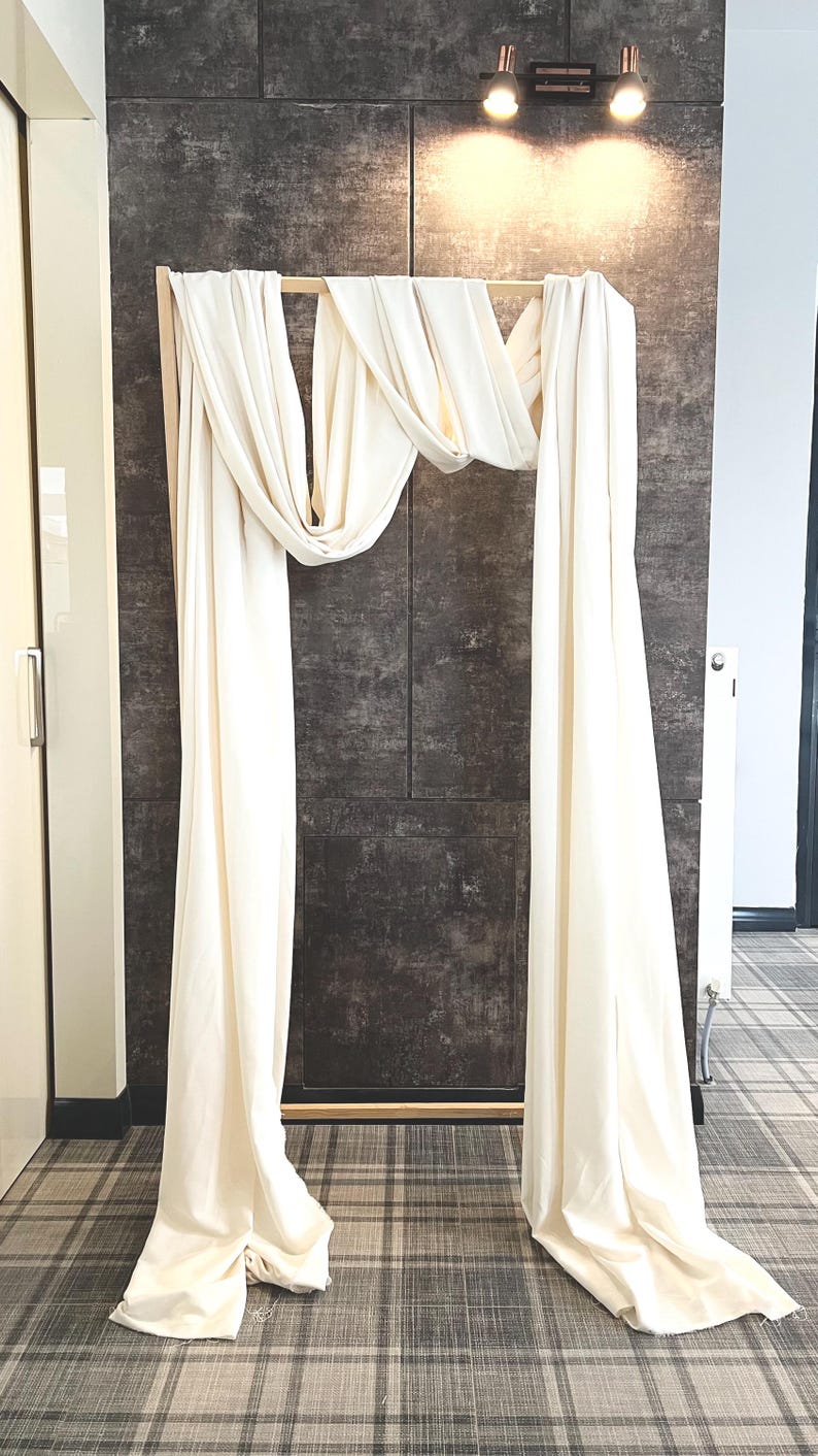 May include: A wooden frame draped with flowing, off-white fabric, creating an archway. The backdrop is a textured, dark gray wall, with a light fixture above. The floor has a plaid pattern.