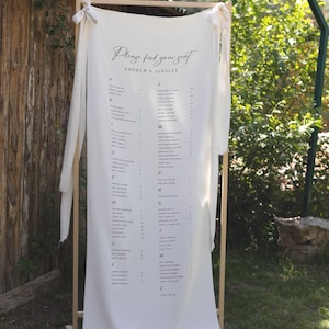 Peut inclure: Un plan de table de mariage sur un cadre en bois. Le tissu blanc affiche le texte "Please find your seat" et les noms "Andrew & Janelle". Les noms des invités sont listés par ordre alphabétique. Le panneau est à l'extérieur.