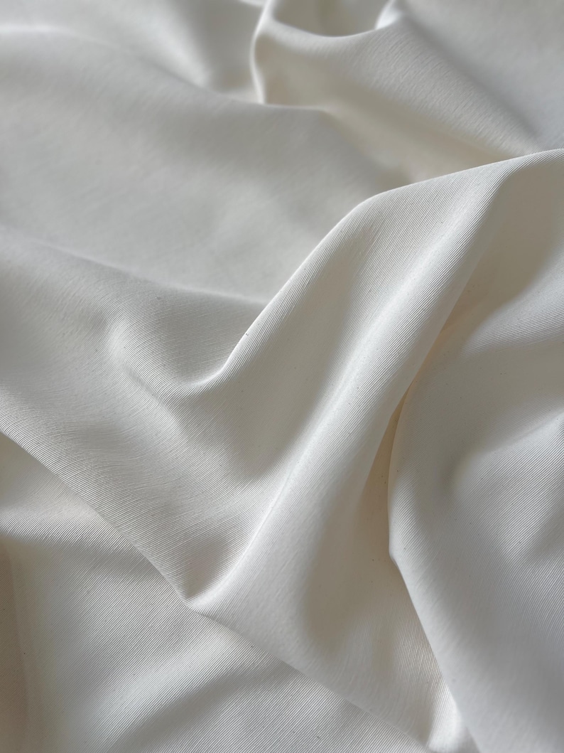 May include: Close-up of off-white fabric with soft folds and subtle texture. The fabric appears to be lightweight and flowing, with gentle shadows creating depth. The material has a smooth, matte finish.