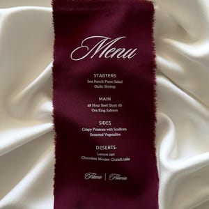 May include: A rectangular deep burgundy fabric menu with frayed edges and white printed text. The menu lists starters, main courses, sides, and desserts, including Sea Ranch Farm Salad, Garlic Shrimp, 48 Hour Beef Short rib, Ora King Salmon, and Lemon tart.