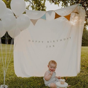 May include: A birthday scene outdoors featuring a child and decorations. A white banner with "HAPPY BIRTHDAY JAX" is visible. White balloons are tied together. The child sits on the grass with a small white stool and a cupcake.