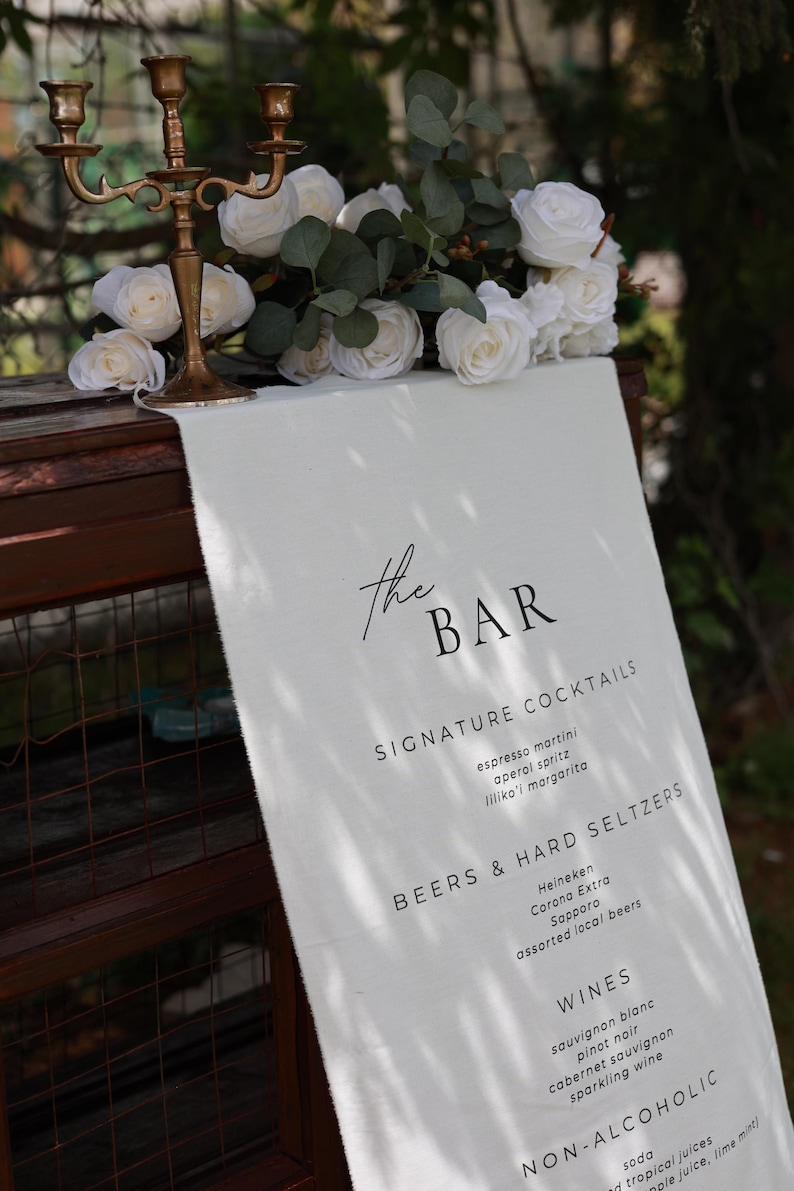 Może przedstawiać: Prezentacja baru z białym lnianym znakiem z napisem "The BAR". Znak wymienia koktajle, piwa, wina i napoje bezalkoholowe. Zabytkowy mosiężny świecznik i białe r&oacute;że zdobią scenę.