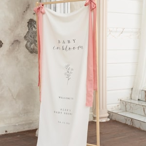 Könnte beinhalten: Ein weißes Stoffbanner mit den Worten "BABY in bloom" in schwarzer Schrift, einer floralen Illustration und Veranstaltungsdetails. Das Banner ist an einem Holzrahmen aufgehängt und mit rosa Bändern verziert.