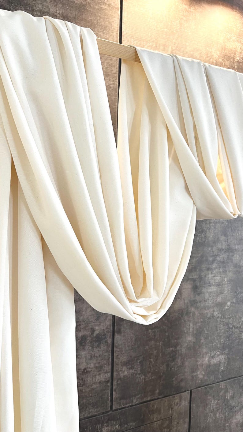 May include: Cream-colored fabric draped over a wooden rod, creating soft folds and shadows. The fabric hangs against a textured, dark gray wall, suggesting a home decor or textile product.