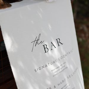 Może przedstawiać: Białe lniane menu barowe z czarnym tekstem, z napisem "the BAR" na g&oacute;rze. Zawiera listę koktajli, piw, hard seltzer&oacute;w i win. Białe r&oacute;że i zieleń w tle.