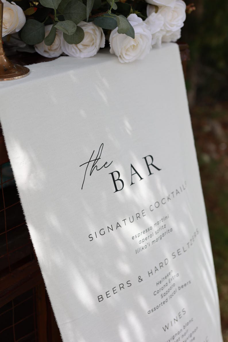Może przedstawiać: Białe lniane menu barowe z czarnym tekstem, z napisem "the BAR" na g&oacute;rze. Zawiera listę koktajli, piw, hard seltzer&oacute;w i win. Białe r&oacute;że i zieleń w tle.