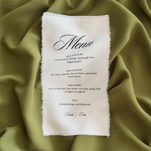 May include: A rectangular white paper menu with frayed edges featuring black printed text including the word Menu, sections for Baslangiclar, Ara Sicaklar, Ana Yemek, Tatli, and the names Irem and Can, placed on a draped olive green fabric.