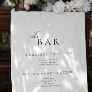 Może przedstawiać: Biały szyld menu barowego z napisem "the BAR" w eleganckiej czcionce. Menu zawiera koktajle firmowe, piwa, hard seltzery i wina. Białe r&oacute;że i zieleń zdobią g&oacute;rną część szyldu.