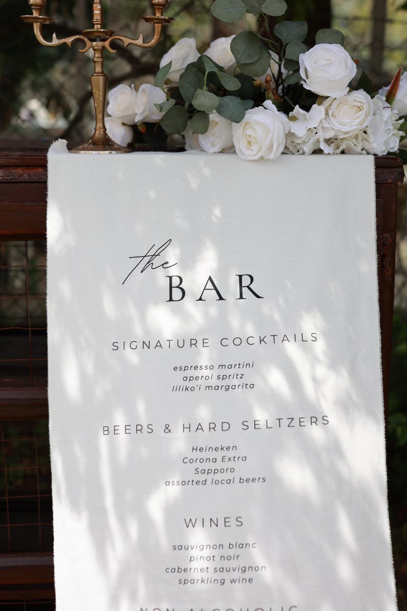 Może przedstawiać: Biały szyld menu barowego z napisem "the BAR" w eleganckiej czcionce. Menu zawiera koktajle firmowe, piwa, hard seltzery i wina. Białe r&oacute;że i zieleń zdobią g&oacute;rną część szyldu.
