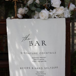 Może przedstawiać: Biały szyld z napisem "the BAR" oraz listą koktajli firmowych, piw, hard seltzer&oacute;w i win. Szyld jest ozdobiony białymi r&oacute;żami i zielenią oraz złotym świecznikiem.